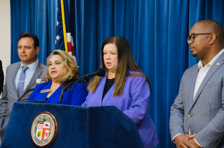 City Attorney Hydee Feldstein Soto at a podium, speaking into a microphone, flanked by two men and another women - mayors and leaders from other local cities.