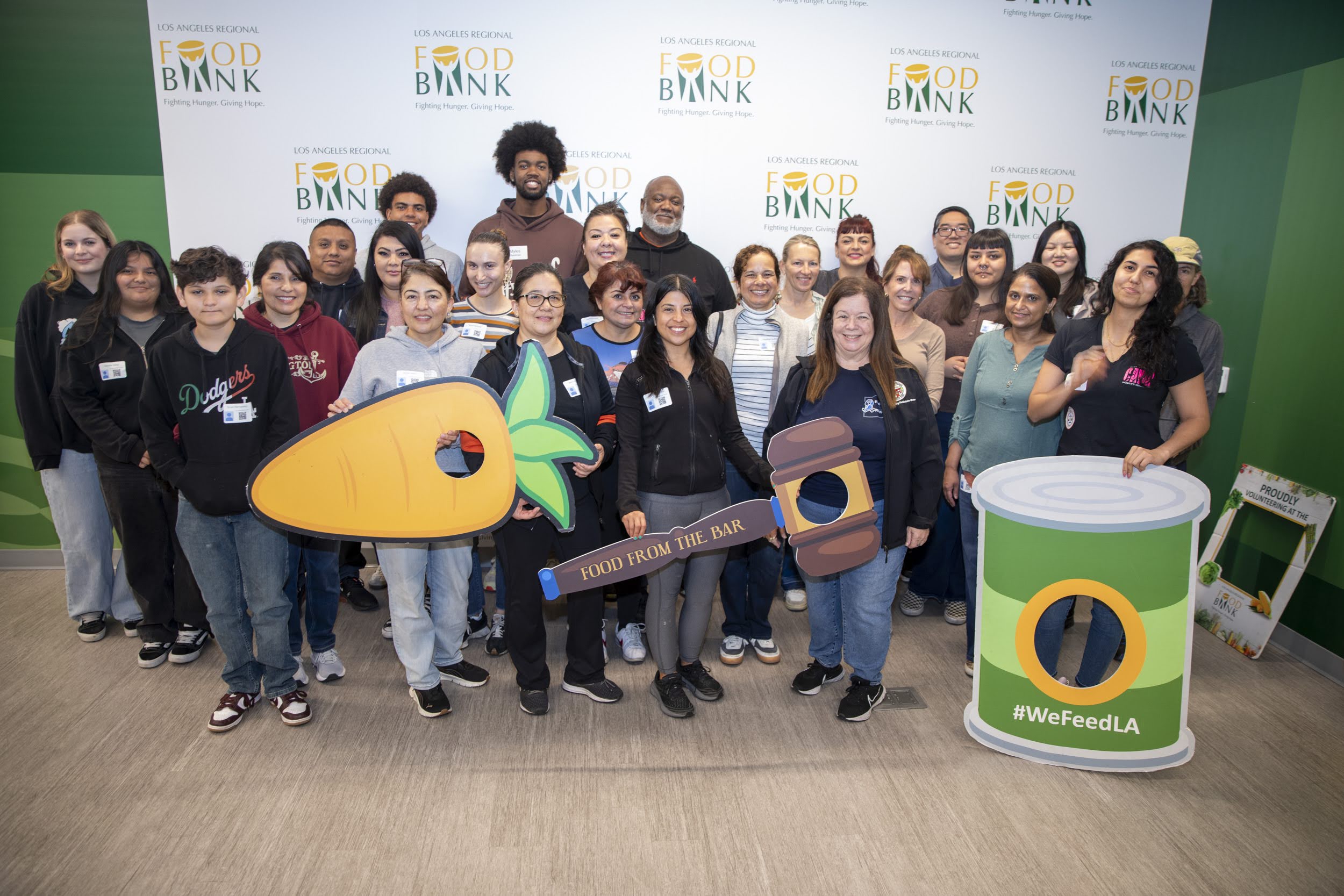 Los Angeles City Attorney Hydee Feldstein Soto standing in front of a group of about 30 people of all ages and backgrounds at a volunteer food bank event. Behind them is a white wall with the logo for the Los Angeles Regional Food Bank on it.