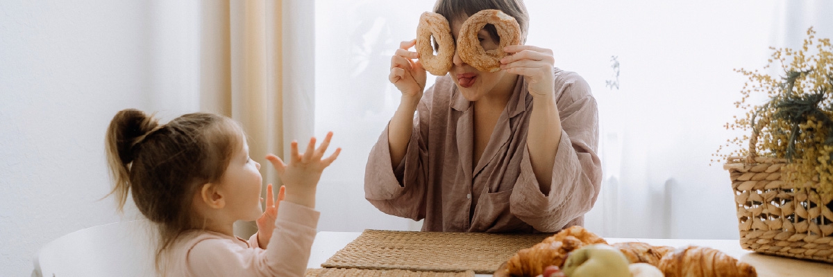 A woman holds two halves of a bagel over her eyes, looking through the holes, as her toddler daughter laughs and throws up her hands.