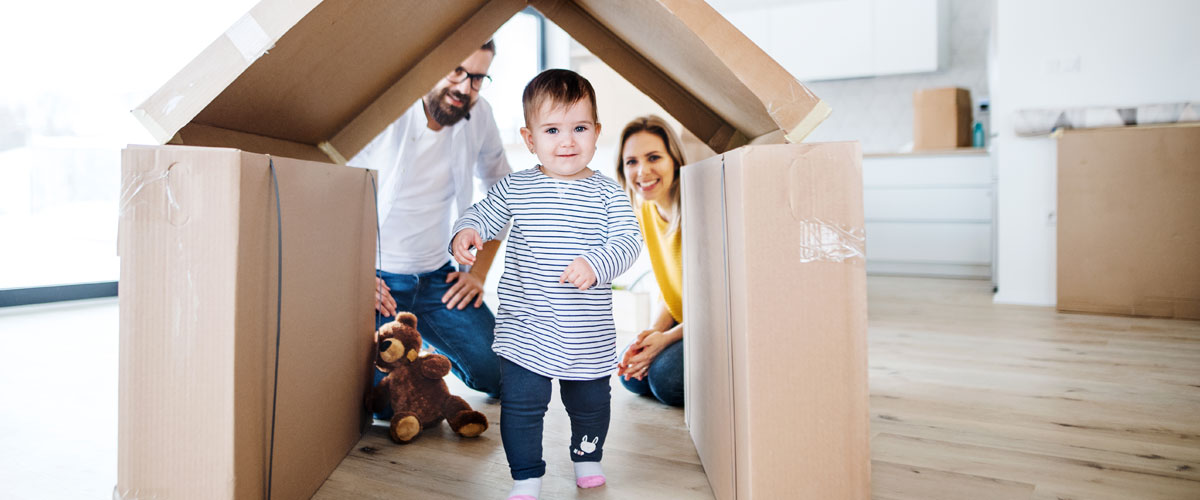 Toddler walking through a cardboard house while two adults watch in background.