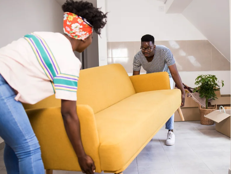 Black couple carrying a yellow couch into a room. Moving boxes are on the floor.
