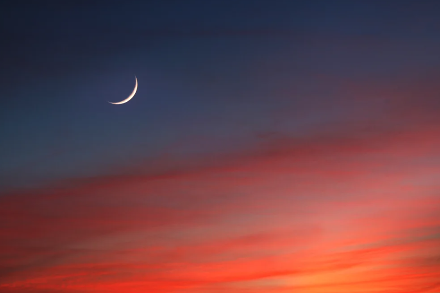 Photograph of a sliver of moon above the pinks and oranges of a sunset