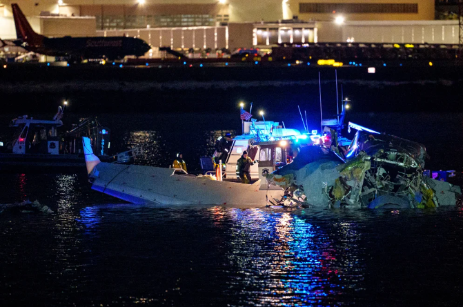 Night-time photo showing emergency response workers inspecting airplane wreckage in the Potomac River, with Reagan Washington Airport buildings in background and ambient aqua and purple-y light reflecting on surface of river water
