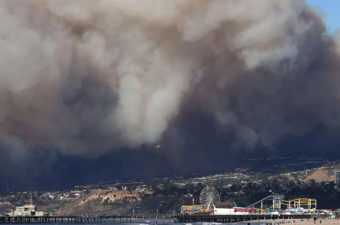 Photo of the Palisades Fire in Los Angeles on Jan. 7, 2025, shows tan and beige smoke roiling over a blackened lower sky with coastal cliffs and nearby residences dotting the landscape. The foreground includes a ferris wheel on a boardwalk.