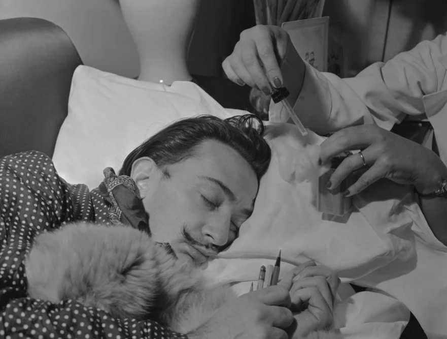 Photograph of Salvador Dali sleeping on a pillow while a woman hovers over his eyes with a dropper