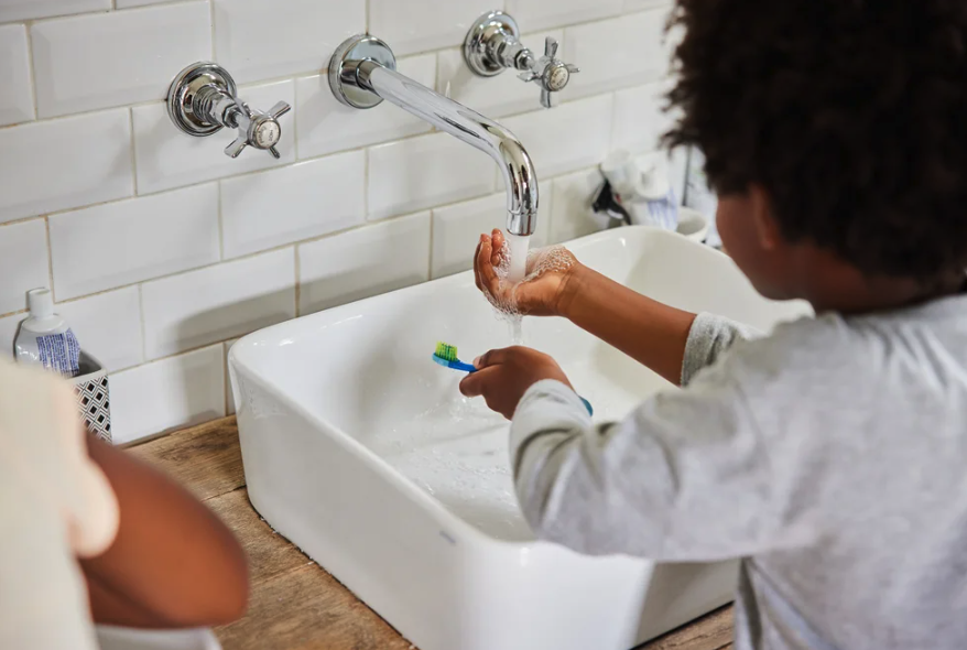 A kid holds his hand under a running faucet. The other hand holds a toothbrush.