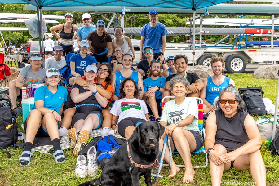 Twenty-one people and one Black Labrador dog sit on grass under a blue tent. Rowing shells on trailers are in the background. 
