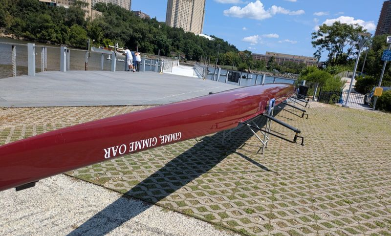 A red eight-oared rowing shell sitting in slings, with the name "Gimme, Gimme Oar" on display.