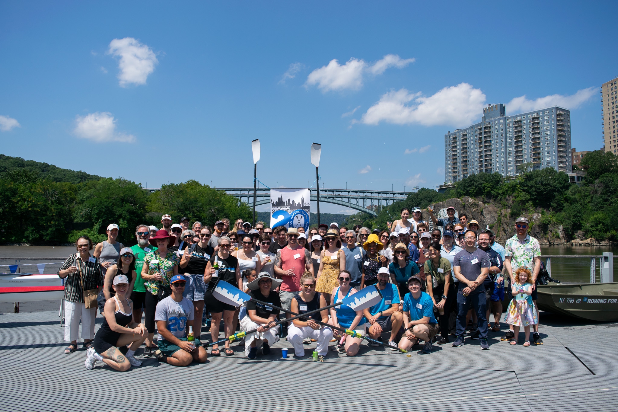 Group photo of the 20th Anniversary party attendees with the river and bridge in the background, a 20th anniversary banner, and some HRCR oars. 