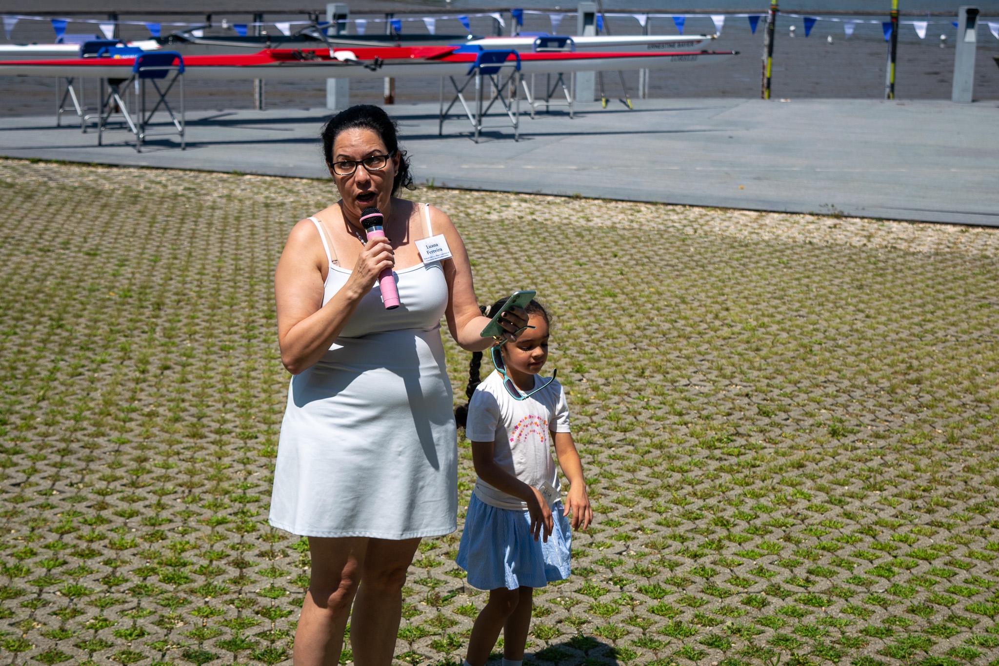 Luana Y. Ferreira holding a microphone next to a small child who is her niece. The rowing shells being christened are in the far background. 