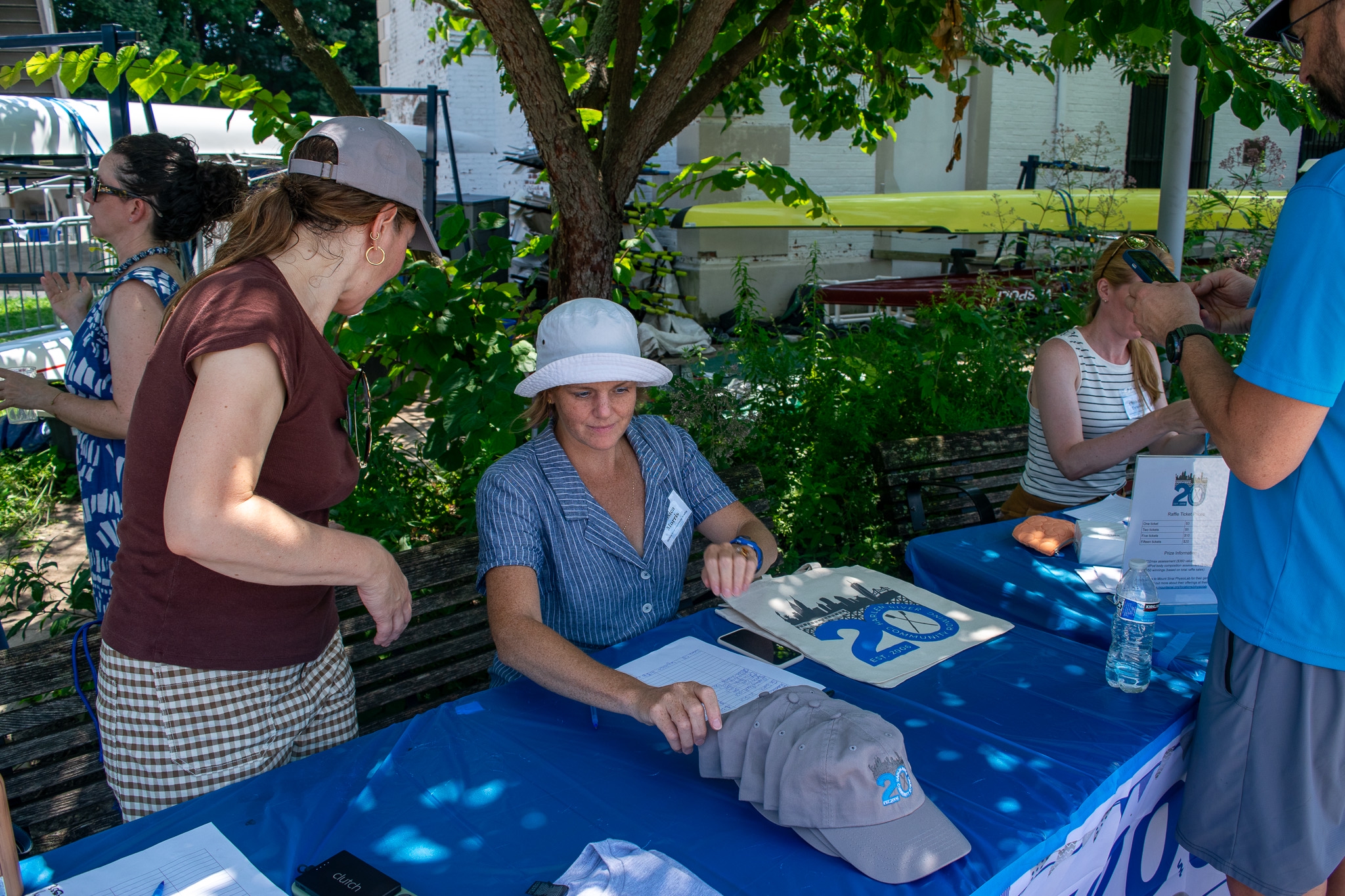 Three people sit behind tables containing hats, t-shirts, and totes for sale, as well a raffle tickets. One person is standing in front a table. 