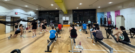 An image from the back of a gym, looking toward eight people on ergometers and approximately eight people engaging in circuit training.