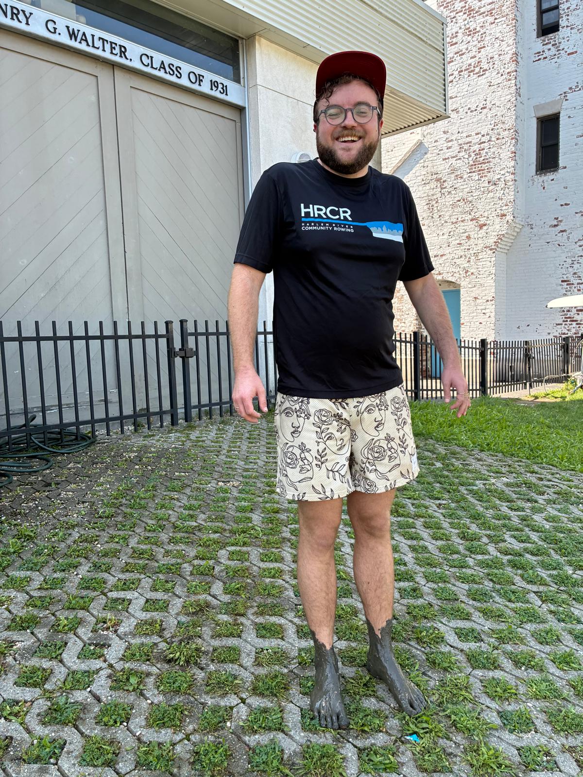 HRCR rower Calvin Harrison standing in front of the Columbia University boathouse. He is wearing a black HRCR t-shirt, is smiling, and has muddy feet. 