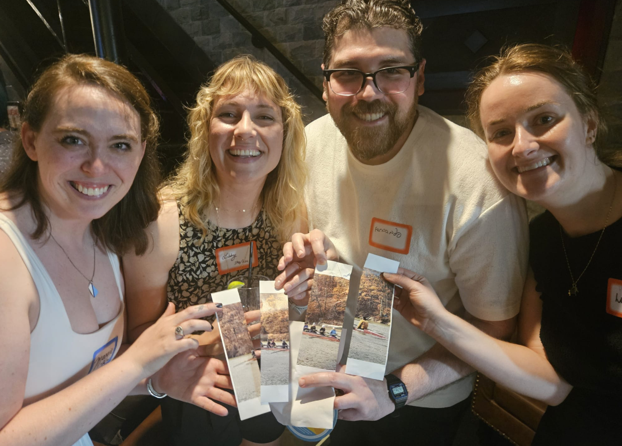 Four people pose for a photo, each holding one quarter of a picture of a boat on a river. The four quarters fit together to form one complete photo.