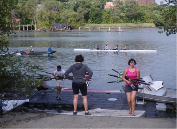 Multiple rowers standing by a dock, some carrying oars, with a four-person shell and a coaching launch in the water in the background.