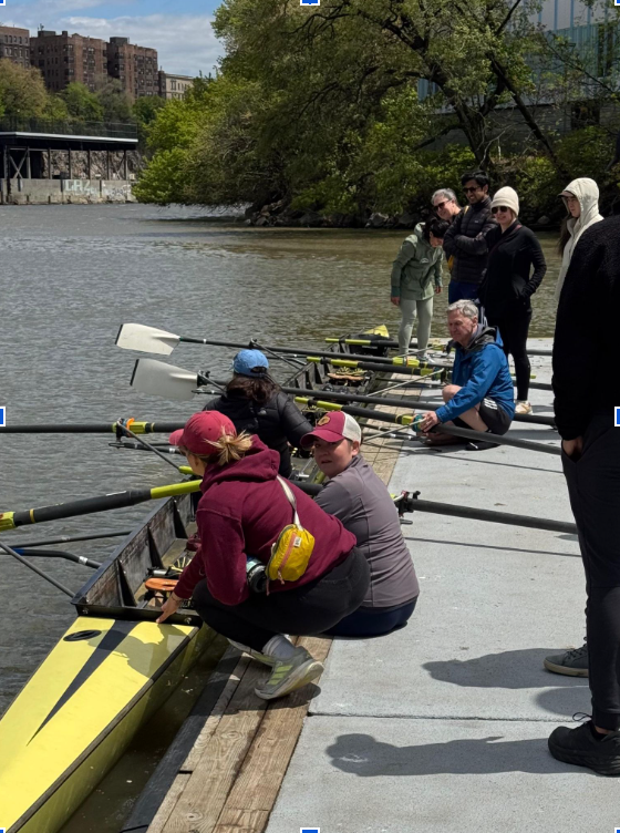 Ten people stand or squat surrounding an eight-oared rowing shell that is resting on a dock.