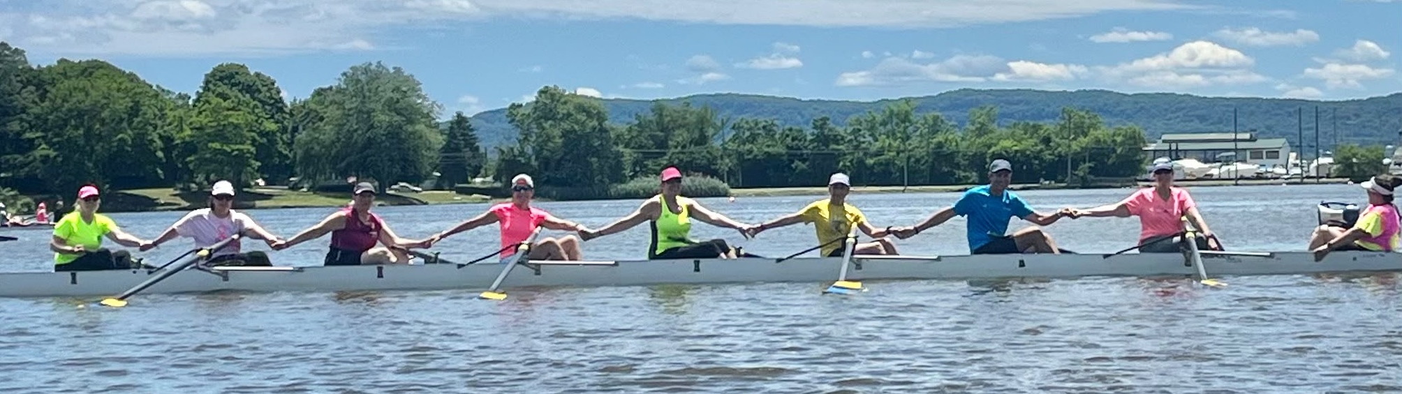Eight rowers and a coxwain in a rowing shell on the water. The rowers are holding hands with the people behind and in front of them. 