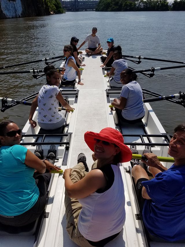 Eight people sit two-by-two in rowing seats in a barge. Two other people sit atop the barge on each end.