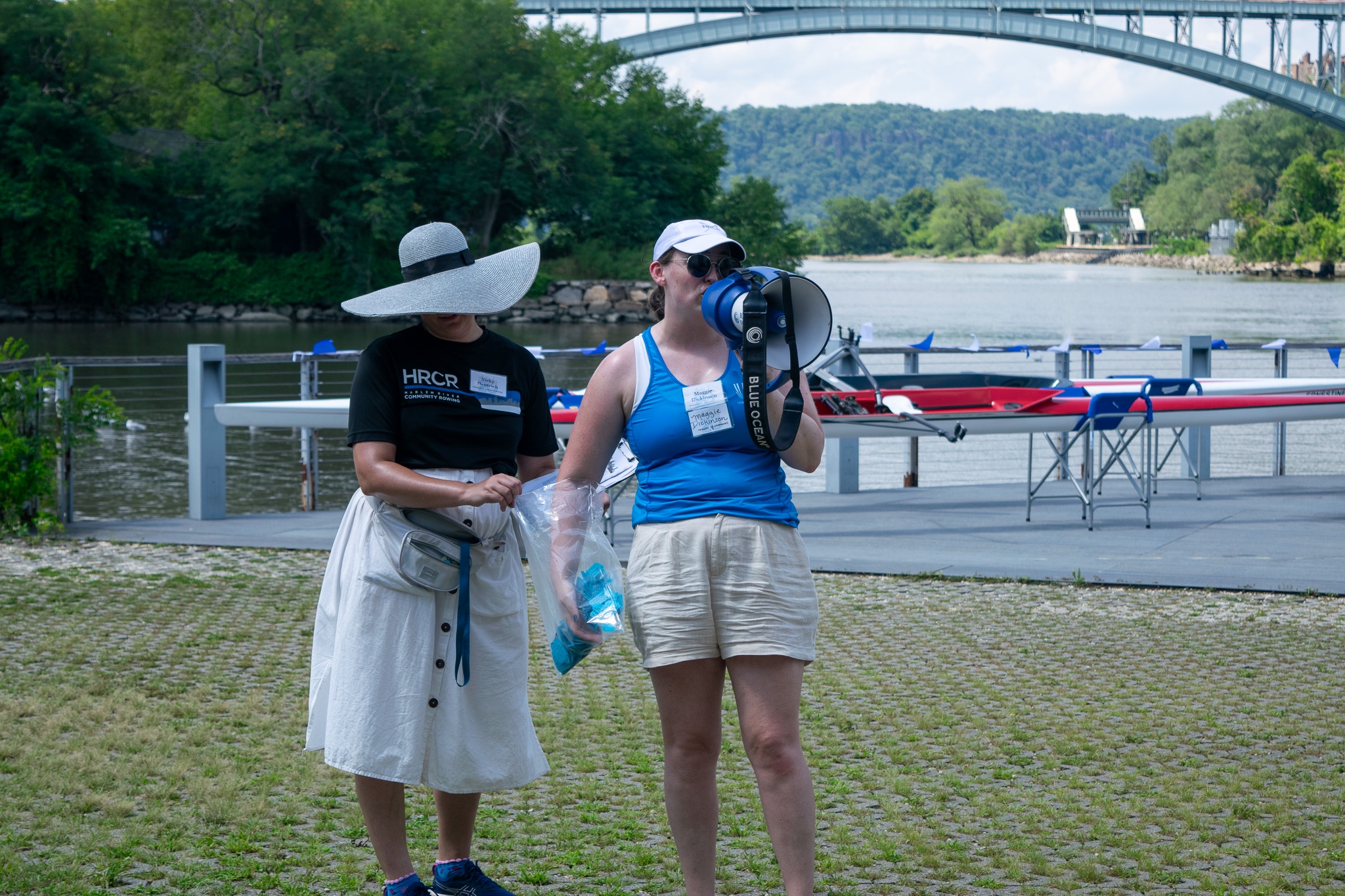 Nicky is wearing a black HRCR t-shirt and a big white hat. Maggie is wearing a blue tank top and holds a megaphone. Maggie is drawing a ticket from the raffle bag, which Nicky is holding. 