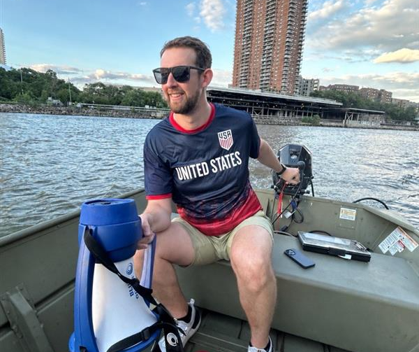 A man (Coach Matt) is holding a megaphone and driving an outboard motorboat on the Harlem River. 