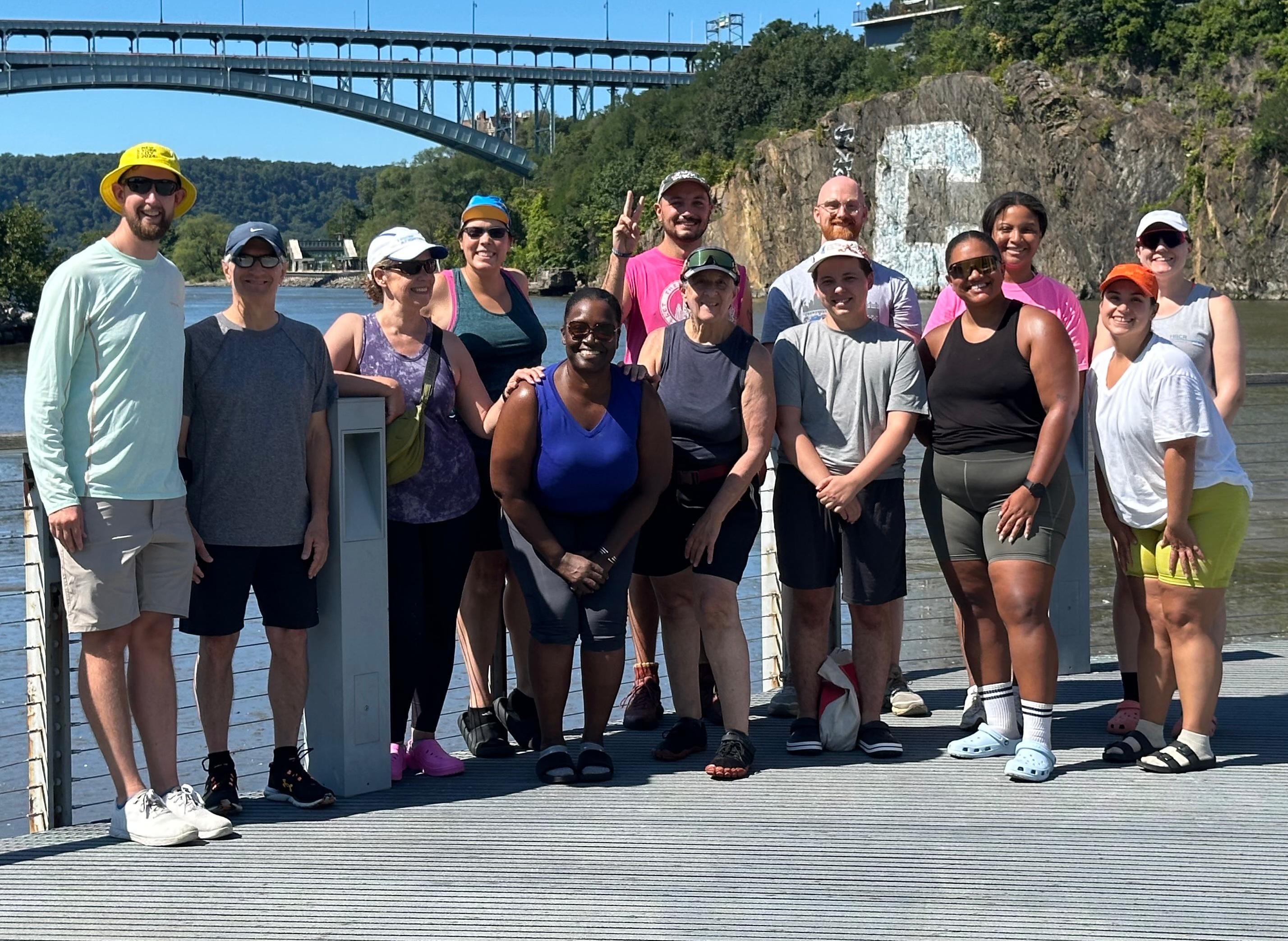 Thirteen people standing in front of the Harlem river with a bridge in the background. 