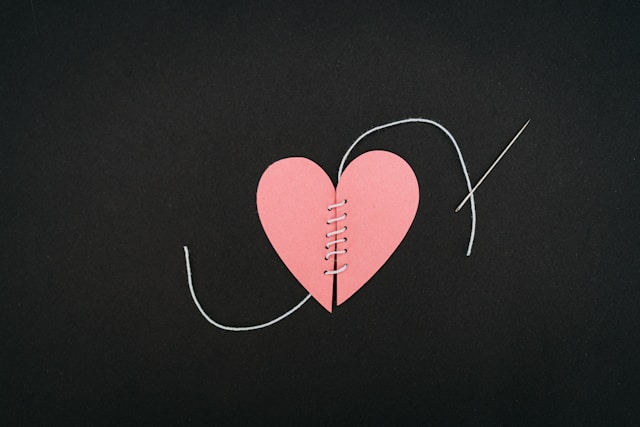 On a black background is a pink paper heart in two and stitched back together with white thread and needle.