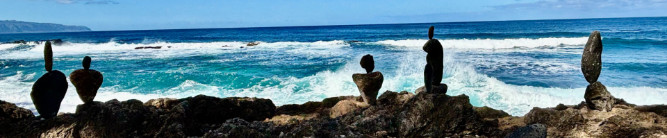 Blue sky and blue ocean, with waves crashing in the distance and in the foreground an outcropping of rock, with cairns built atop.