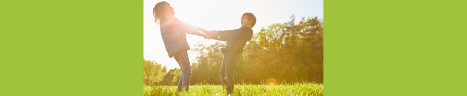 Two children holding hands and spinning in a sunlit grassy field with trees in the background.
