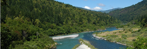 Two rivers converge into one through a lush green valley with tree-covered mountains and pebbled shores on either side under a bright blue sky with scattered clouds.
