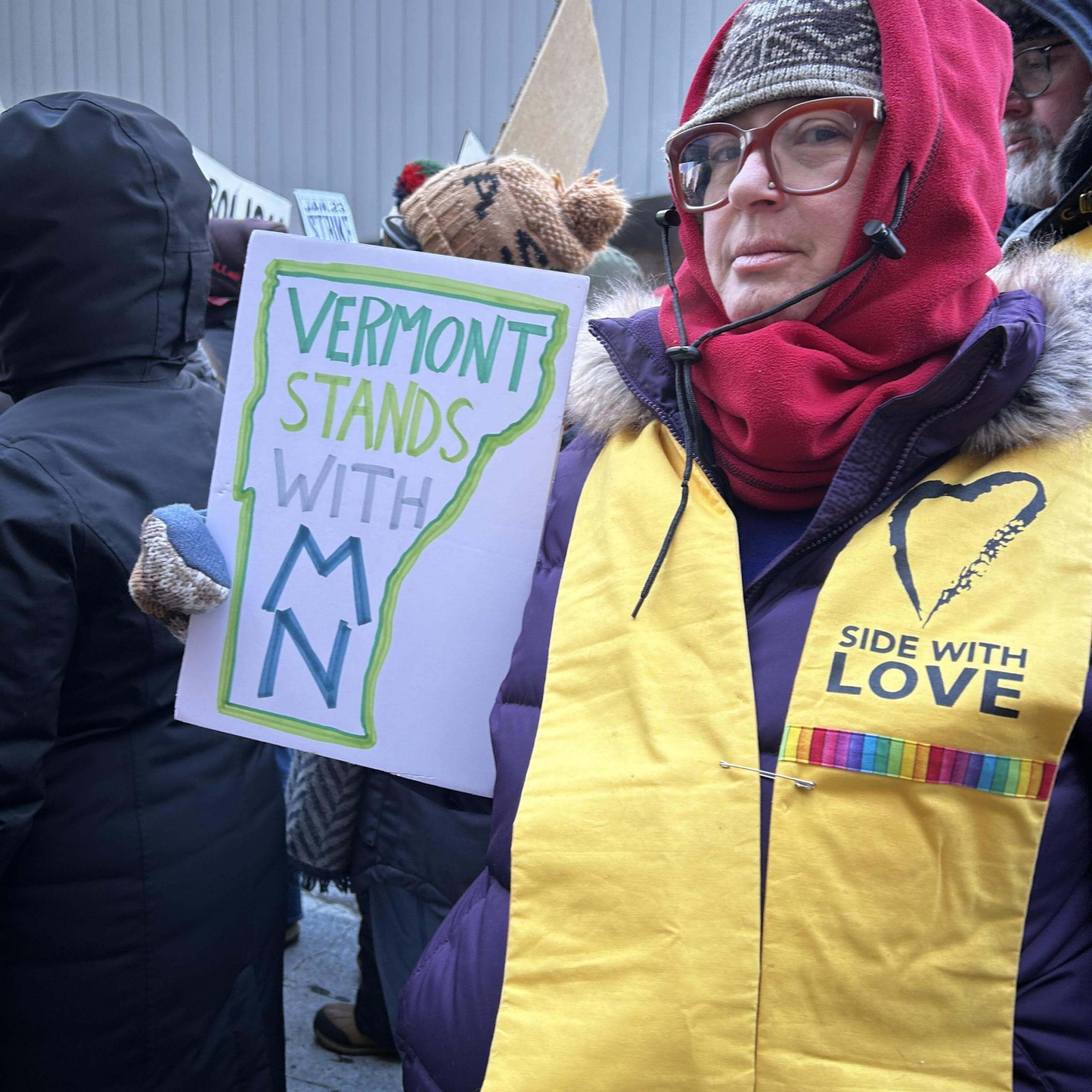 Rev. Karen G. Johnston, senior minister of First UU Society of Burlington, Vermont, holds a sign that says Vermont stands with MN. Johnston is bundled up to stay warm and wearing a yellow Side with Love stole. 