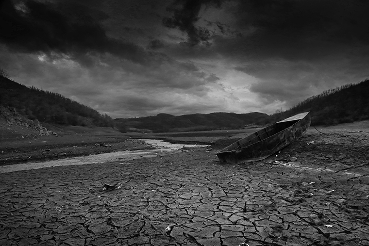 A river with hills in the background surrounded by dry, cracked earth with a johnboat perched on an incline on the right.