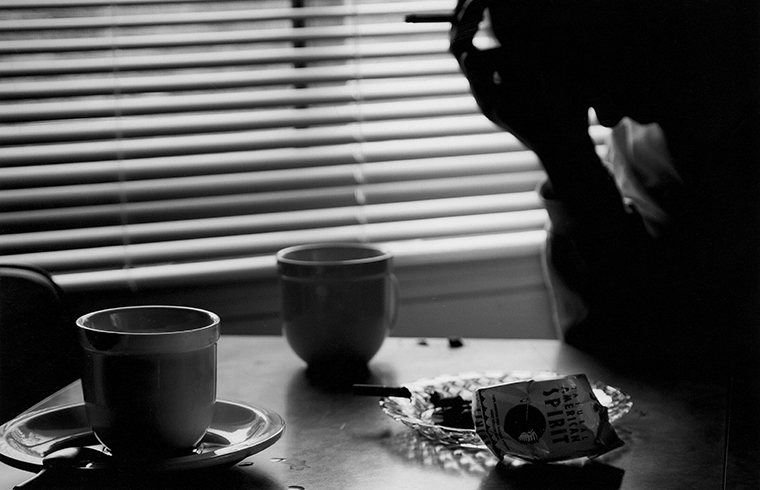 A person sitting at a table in a diner has their elbow on the table and is resting their hand holding a cigarette between their fingers on their forehead. The table has two coffee cups and an ashtray on it.